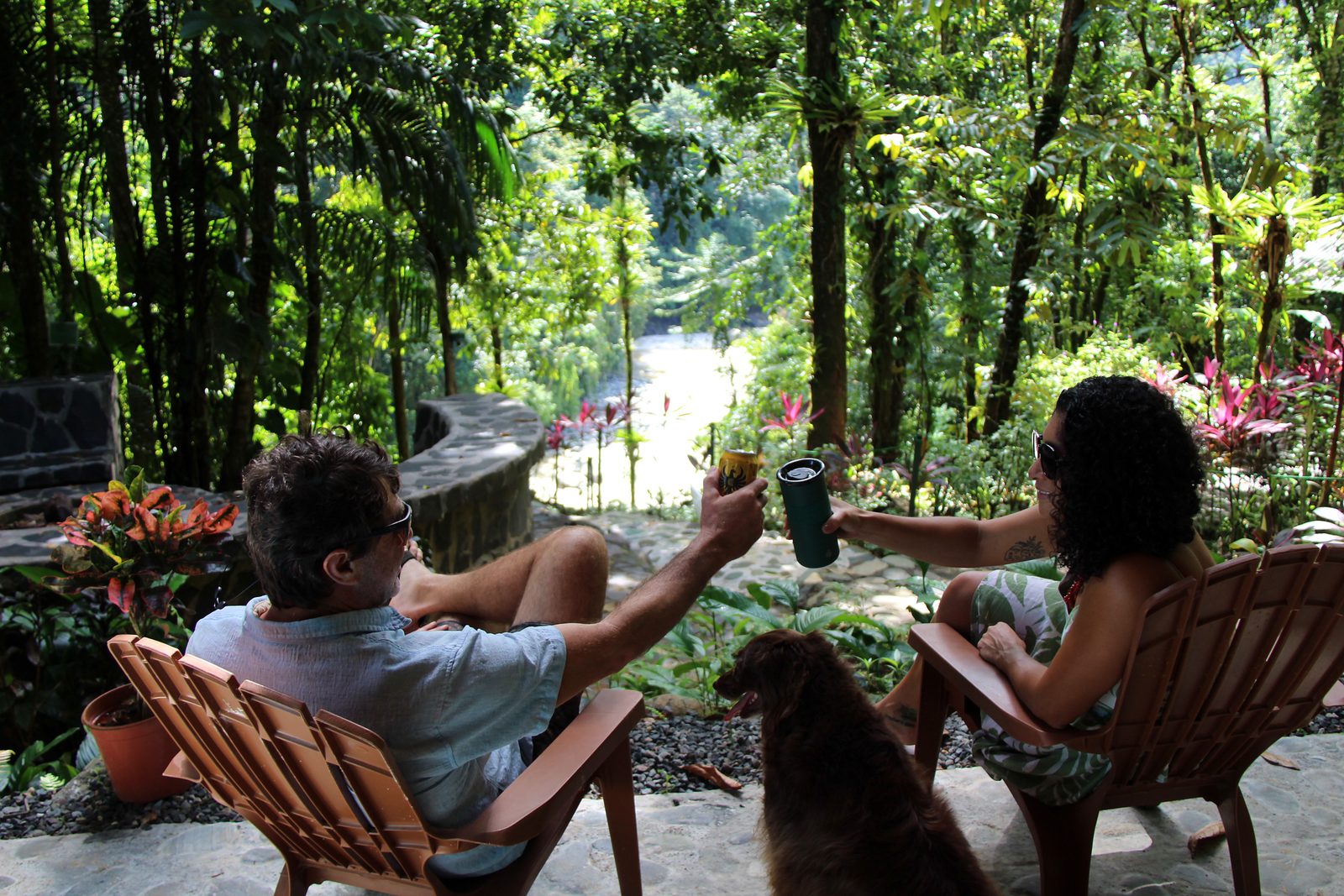 A couple toasting in wooden chairs on the terrace, the dog beside them